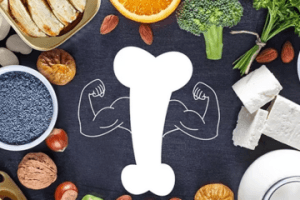 Illustration of a strong bone with flexing arms surrounded by healthy foods that support bone health, including nuts, seeds, tofu, broccoli, dairy, leafy greens, figs, and orange slices.
