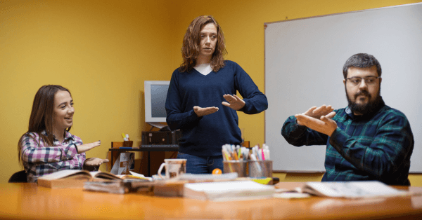 Three adults in a classroom setting practice Auslan together; one woman stands while two others seated at a table follow her signs.