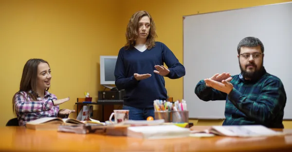 Auslan Cert II 1 Three adults in a classroom setting practice Auslan together; one woman stands while two others seated at a table follow her signs.