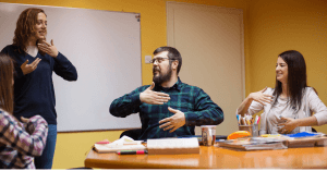 A group of four adults in a classroom use Auslan, smiling and interacting as they practice australian sign language around a table.