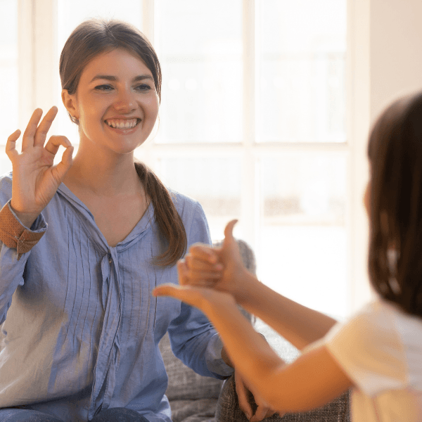 A smiling young woman uses sign language with a child in a bright, comfortable room.