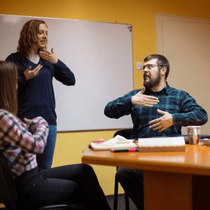 Three adults in a classroom setting practice sign language together in front of a whiteboard.