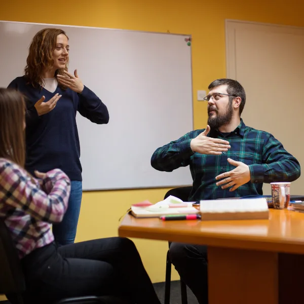 Learn Auslan 3 Three adults in a classroom setting practice sign language together in front of a whiteboard.