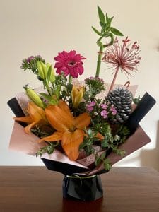 Colourful floral arrangement featuring orange lilies, a pink gerbera, small purple flowers, greenery, lucky bamboo and a silver pinecone, wrapped in pink and black paper.